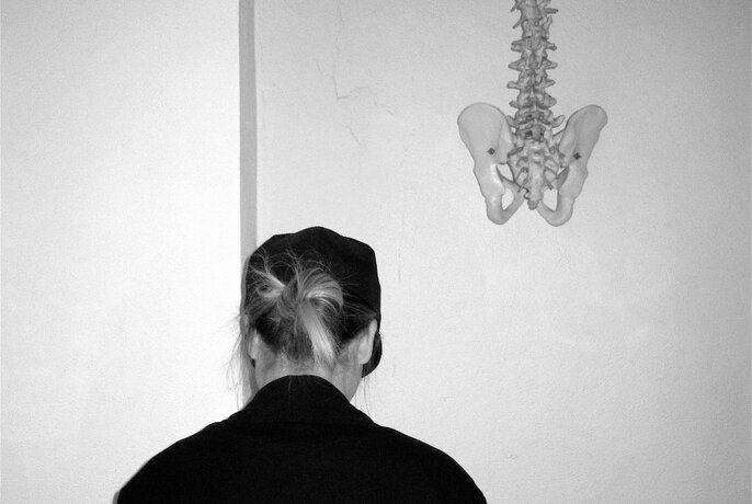 The back of a models head wearing a black jacket and black cap with bun pulled through, there is a spine hanging in the background