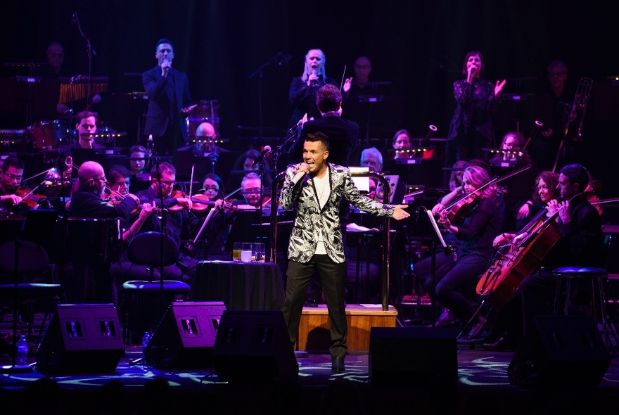 Singer Anthony Callea singing on stage with a symphony orchestra behind him. 