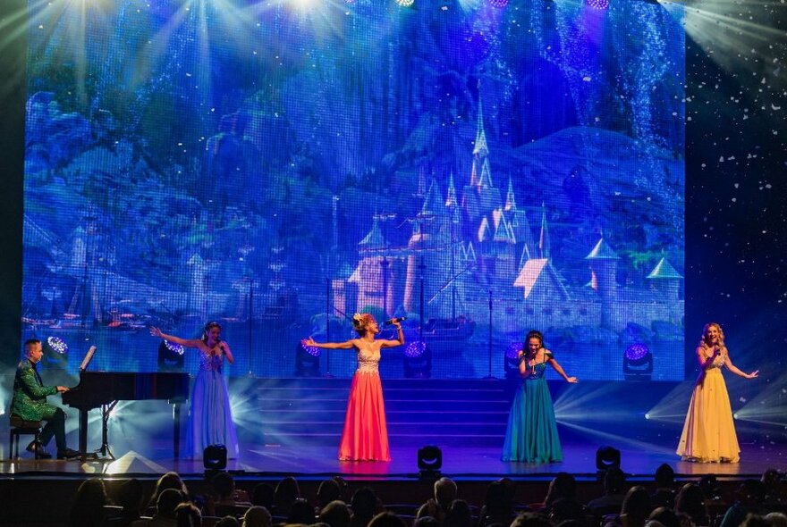 Four women in different coloured dresses and a pianist on stage with a fairy castle appearing in the blue background. 