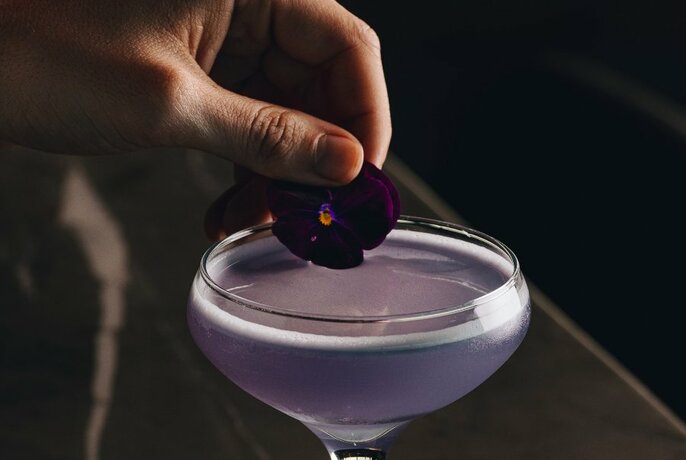 Martini-shaped glass filled with a lavender coloured beverage, with a hand placing a floral garish on the top of the glass.