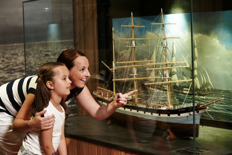A young child and adult looking and pointing at a model of a ship in a maritime museum. 