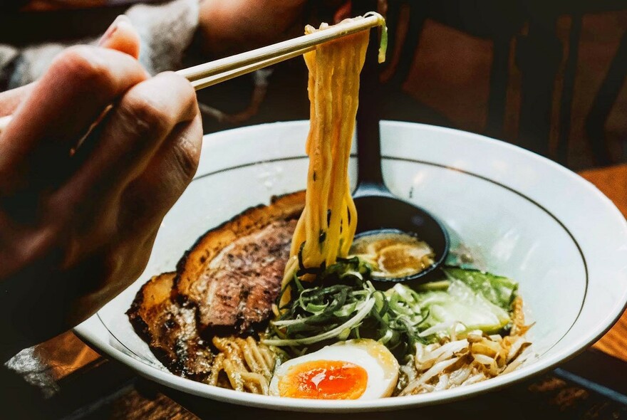 Hand holding chopsticks over a bowl of ramen with egg and vegetables.
