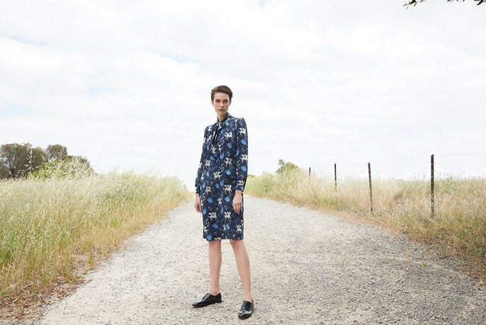 Model wearing a blue patterned dress, standing on an unmade road beside a field.