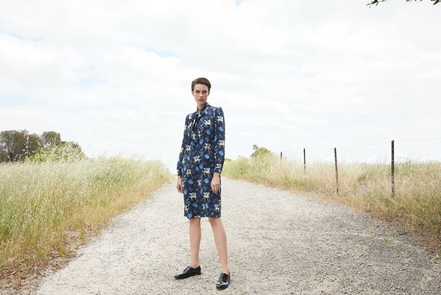 Model wearing a blue patterned dress, standing on an unmade road beside a field.