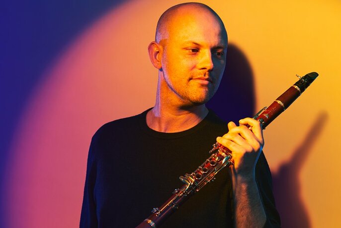 Musician Nicolas Fleury holds a clarinet while a coloured light casts a glow on him and the background wall.