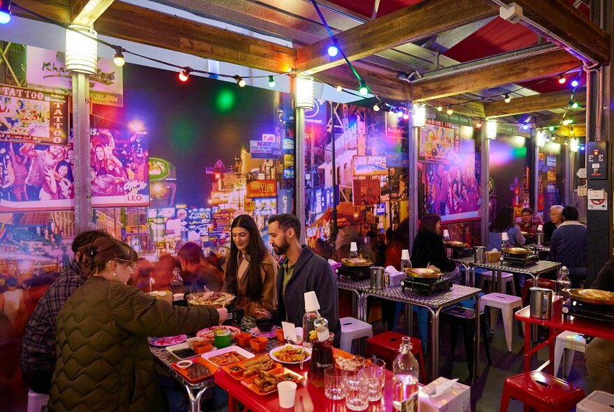 People dining in an outdoor covered laneway restaurant with colourful murals, festoon lights and hotpot dishes.