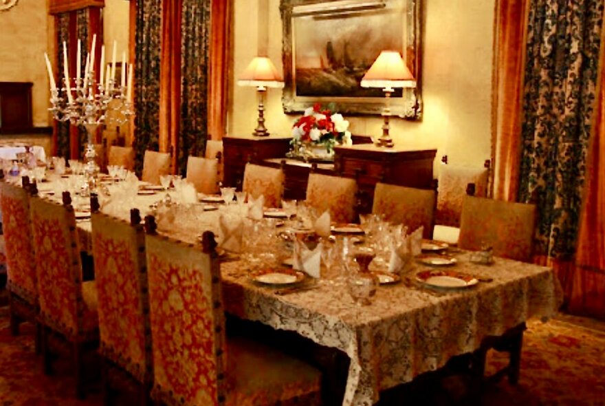 Ornate dining room with a long table set for dinner service, a lace tablecloth, upholstered dining chairs, heavy patterned velvet drapes and soft lamps on a sideboard.