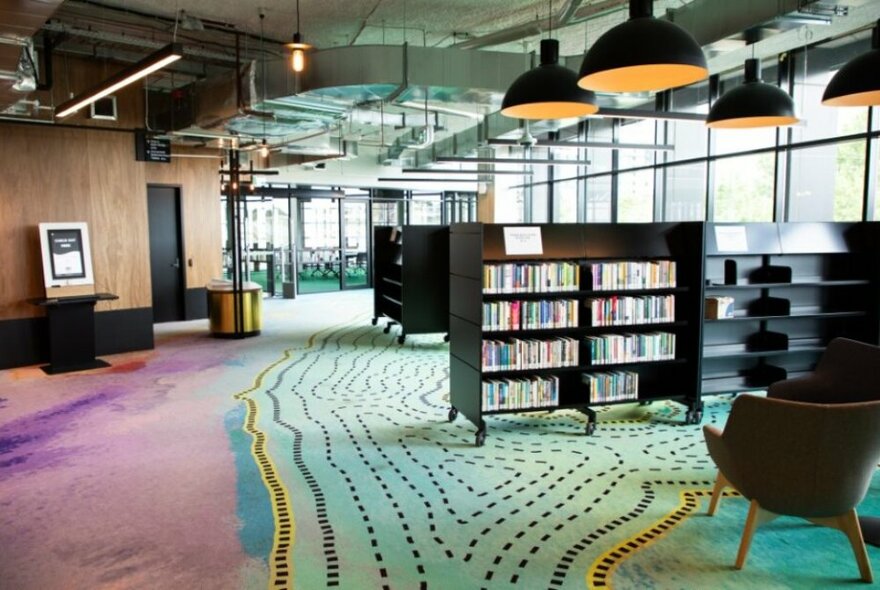 Interior of a large and welcoming library space, with patterned carpet, shelves of books, armchairs and plenty of natural light