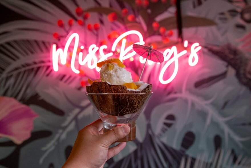 A  hand holding a dessert with umbrella in front of pink neon sign spelling Miss Ping's.