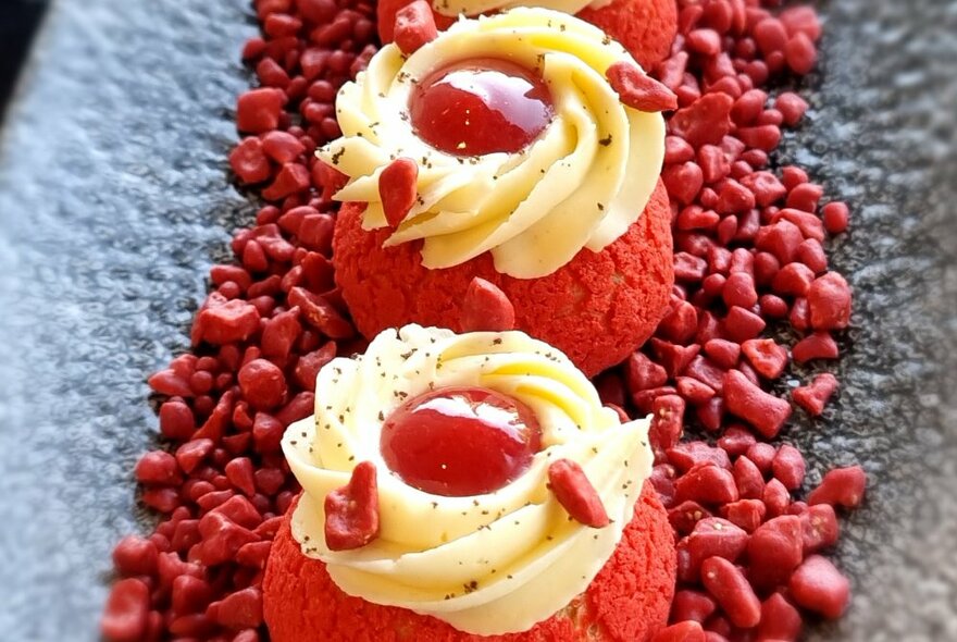 Two red tea-cakes with swirls of cream and jam drops on top on a bed of red crumbs on a dark plate.