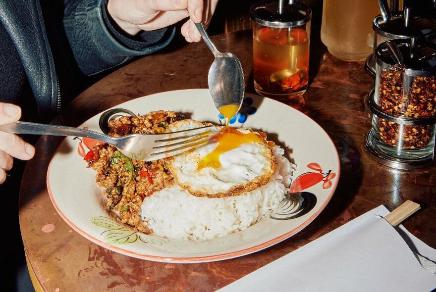Hands just visible holding fork and spoon over egg, rice and vegetables. Condiments in large glasses to right.  