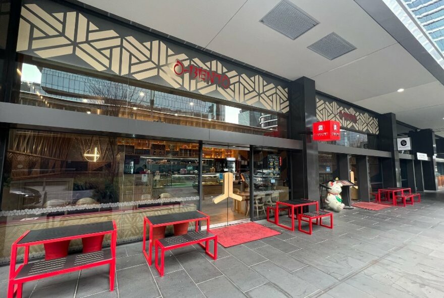 Exterior of a Japanese restaurant with glass front windows and red and black tables and benches on a grey paved footpath.