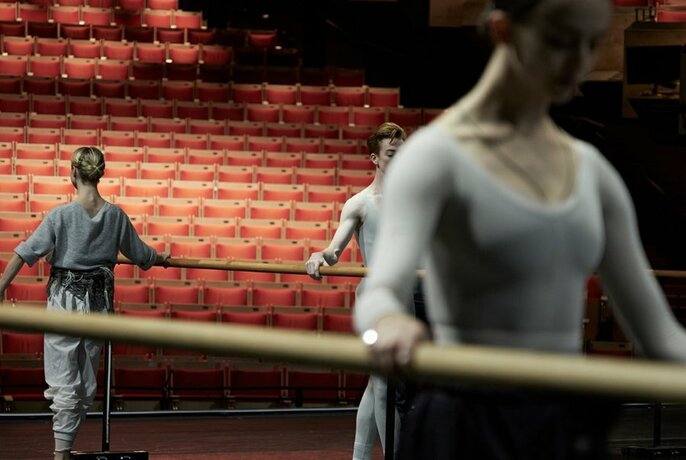 Ballet dancers practising on a stage with barres in front of tiers of seating.