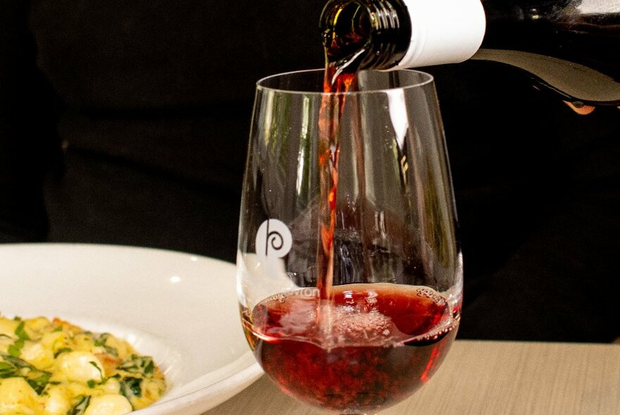 Glass of red wine being poured, next to dish of pasta.