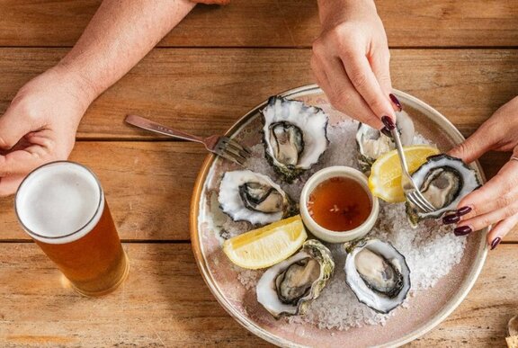 Overhead view of hands hovering over a plate of oysters and selecting one with a small fork, a pot of beer nearby.