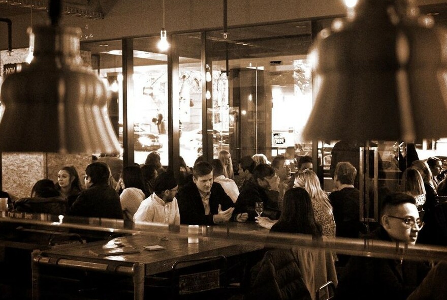 Sepia image of cafe interior with people seated at tables in front of a bar with pendant lights.
