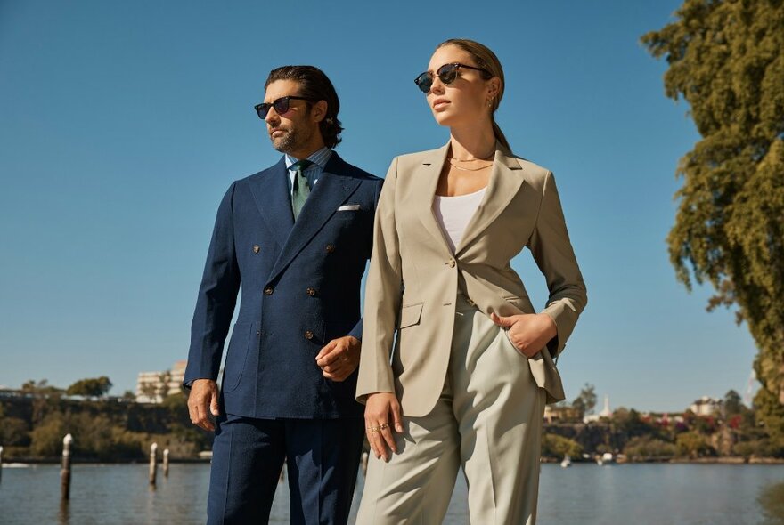 A male model wearing a double breasted navy suit and a female model wearing a loose fitting sand coloured suit, posing outdoors on the edge of a body of water.