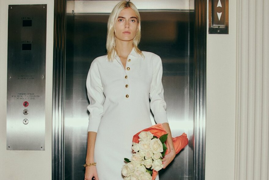 A woman with blond hair modelling a white designer dress, holding a bouquet of flowers in her hand, standing in an open elevator.