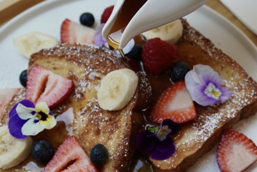 Maple syrup being poured over French toast, and sliced fresh fruit.