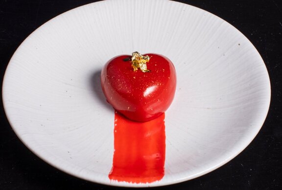 A red heart shaped dessert with gold leaf and a stripe of matching sauce on a plate.