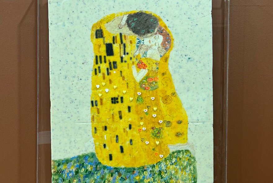 A reproduction of Gustav Klimt's famous The Kiss painting, recreated in chocolate, displayed on a wall behind glass.