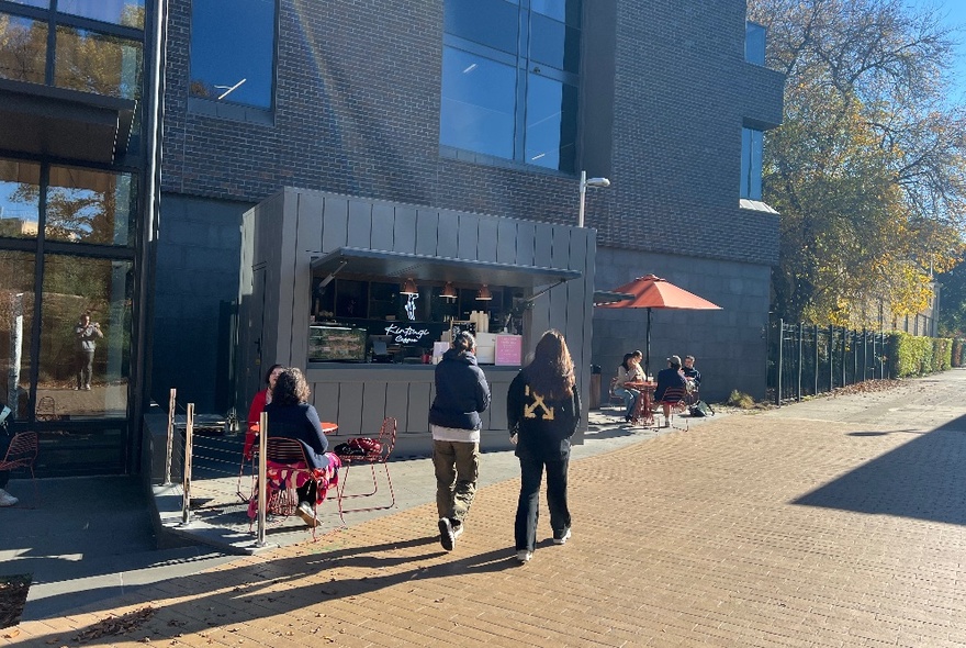 Two people walking past a serving window of a cafe in a black building.