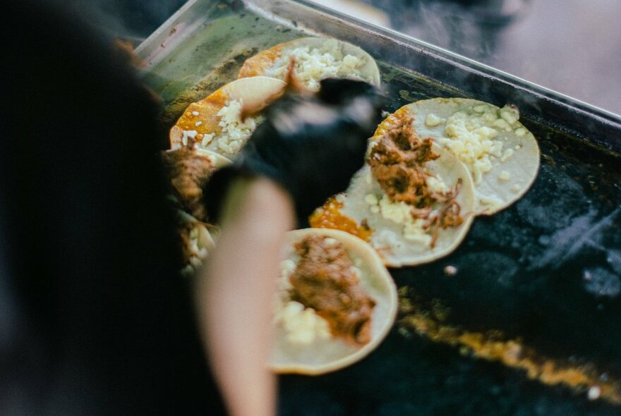 Tacos being prepared by a cook at a hot BBQ grill plate.