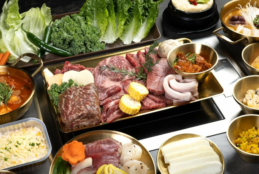 A plate of meats, seafood and vegetables waiting to be cooked on a table-top BBQ grill, with many small bowls of side dishes.