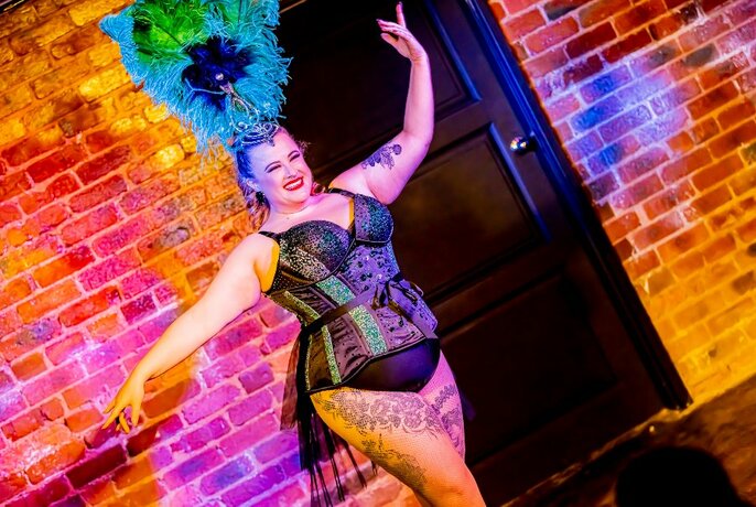 Burlesque performer in a black corset costume, and blue feathered headdress, arms posed on either side of them, on a stage.
