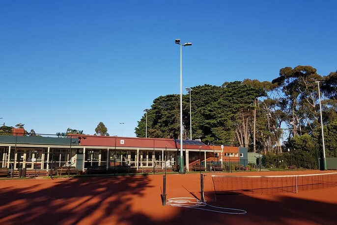 Red clay tennis courts in foreground with Royal Park Tennis Club and large green trees in the background.