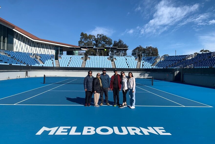 A group of six people posed near the central net on a blue tennis court at Melbourne Park tennis arena, the word MELBOURNE printed on the court in large white upper case letters, with spectator stands, blue sky and trees visible in the background.