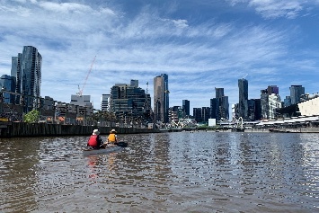 The Yarra River with two people in a canoe visible in the middle ground, and the city skyline in the background.
