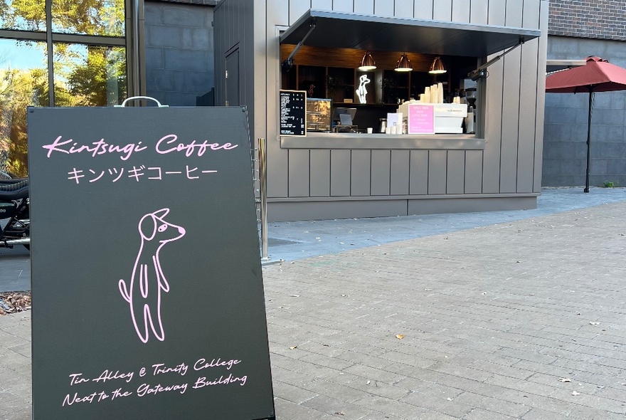 An A-frame sign with a pink line drawing of a dog standing on its hind legs, in front of a serving window of a cafe.