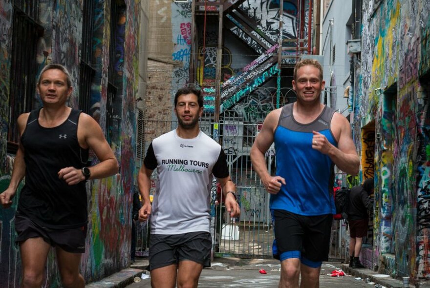 Three men in singlets and shorts running down a laneway with the walls covered in street art.