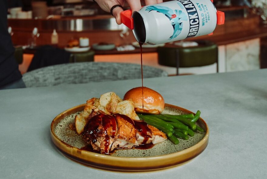 Roast chicken with sides on a brown plate with gravy being poured from a beer can above.