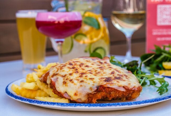 A chicken parmigiana with chips and salad surrounded by drinks. 