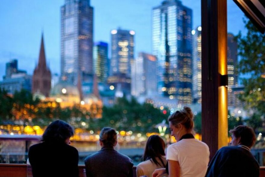 Diners at a riverfront venue with blurred city buildings at twilight in the background.