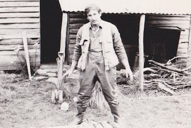 Old black and white photograph of a man in fishing waders and fishing vest standing in front of a log cabin.