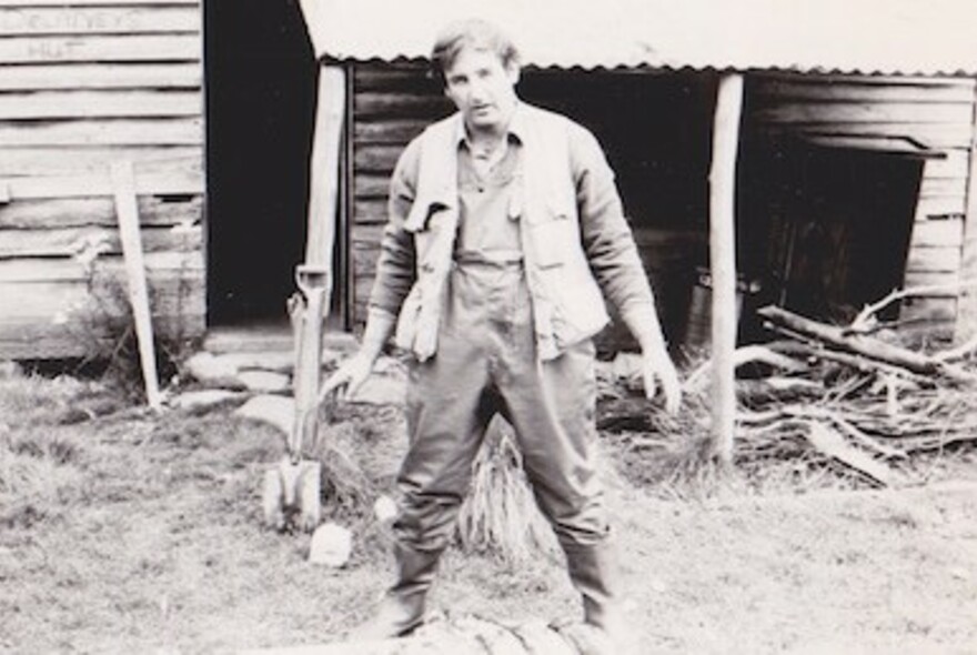 Old black and white photograph of a man in fishing waders and fishing vest standing in front of a log cabin.