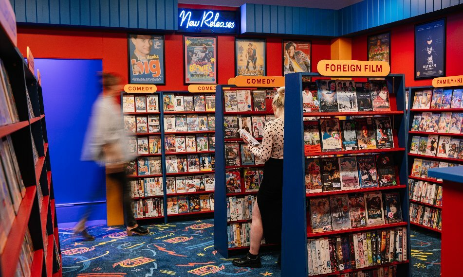 Two people browsing through shelves of movies in a nostalgic exhibit designed to look like a home video rental shop from the 1990s.