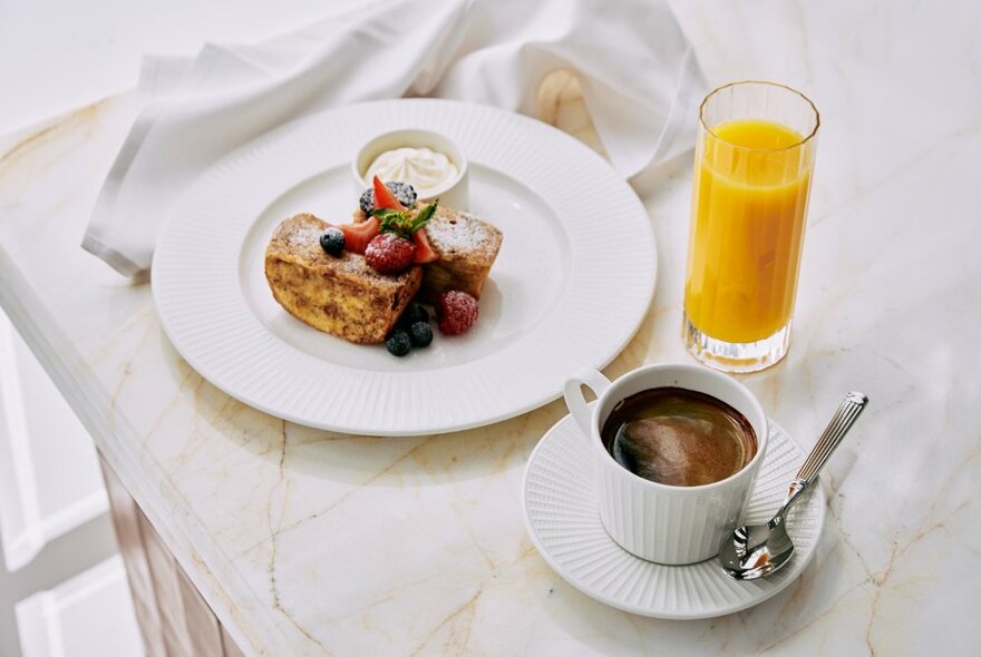 Glass of orange juice, a cup pf coffee and a plate with breakfast pastries and fruit, on a marble table top.