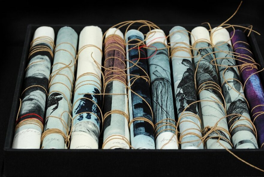 A box containing nine rolled-up etchings, lying side-by-side and held together with thin string, the image in each one facing outwards.