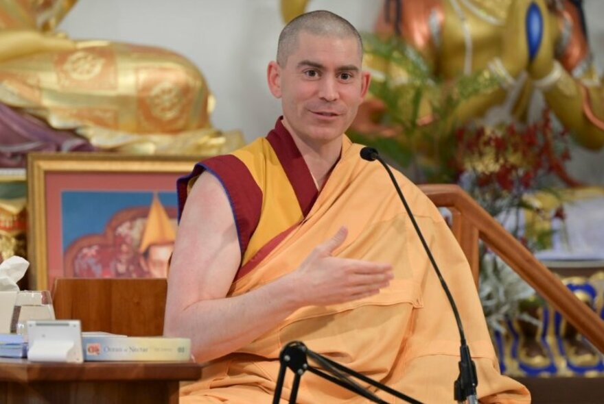Buddhist monk wearing saffron robes seated at a microphone in front of gilded statues.