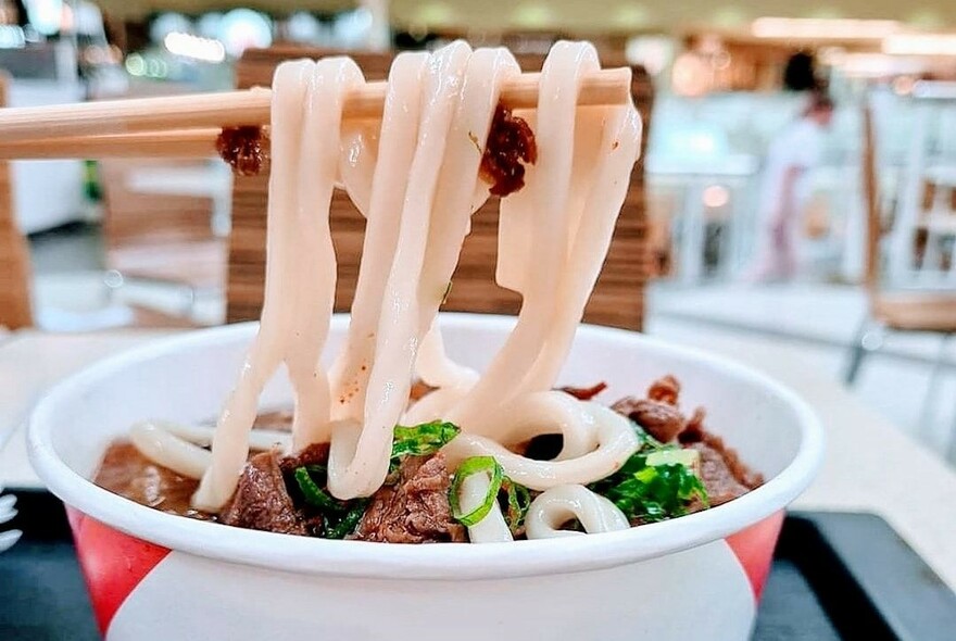 Fat soba noodles hanging off a chopstick over a bowl of soup.