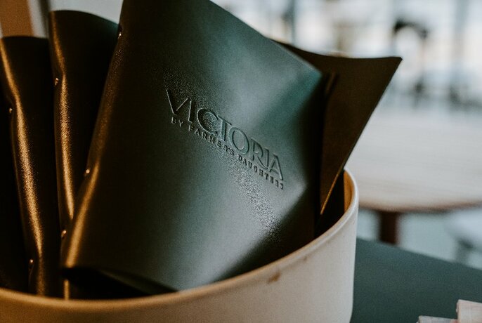 A closeup of a black leather bound menu with the words Victoria By Farmer's Daughters embossed onto the front cover.