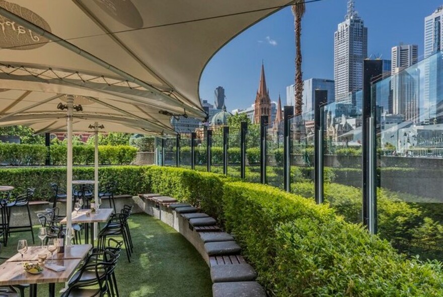Curved patio seating against a clipped hedge next to dining tables under outdoor sails, with city skyscrapers in the background.