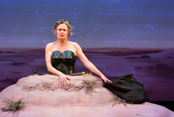Performer Judith Lucy from the waist up, the lower half of her body buried in a small hill, on a stage. 