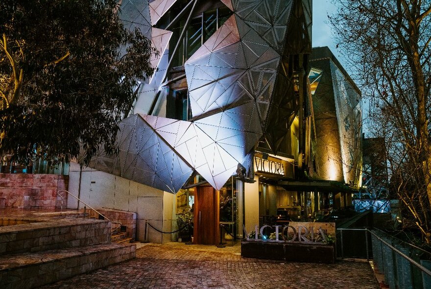 Exterior of a restaurant entrance at Fed Square, showing evening sky and trees and a spotlight shining on the building's distinctive facade.