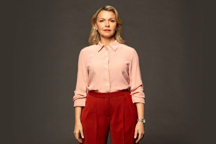 Actress Justine Clarke standing with arms by her side, wearing red trousers and a pale pink shirt, looking directly at the viewer.
