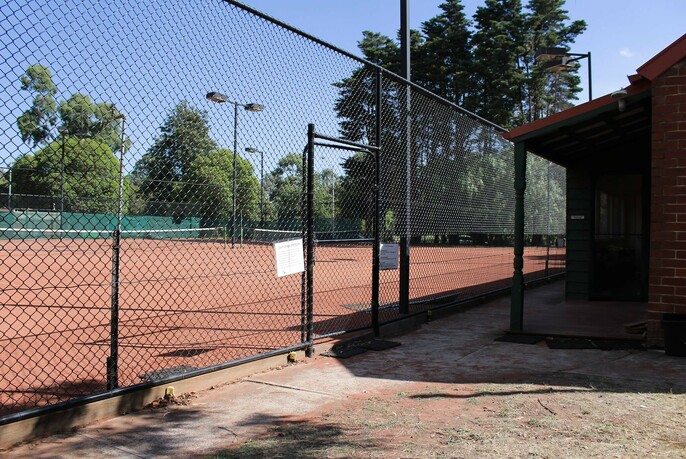 Two fenced tennis courts with trees and blue sky in the background.